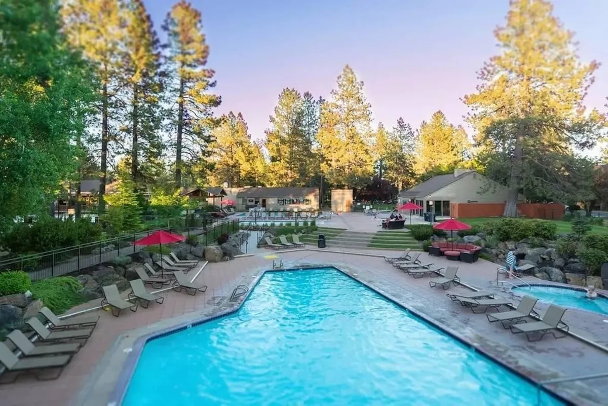 A vibrant exterior shot of the resort's swimming pool, surrounded by sun loungers and tall trees. Patio umbrellas provide shaded seating, and the lush outdoor area invites guests to relax and socialize. The scene is framed by the natural beauty of Central Oregon, hinting at the abundant outdoor experiences available nearby.