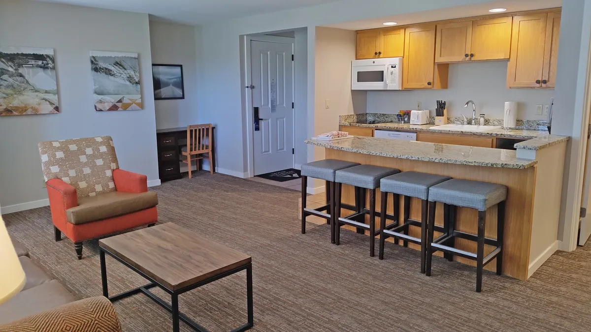 The image shows a warm, inviting interior of a condo with a modern kitchen area featuring sleek countertops and bar stools, an armchair positioned for relaxation, and a cozy coffee table perfect for gathering. The scene suggests a space ideal for cooking, entertaining, and unwinding with friends or family.