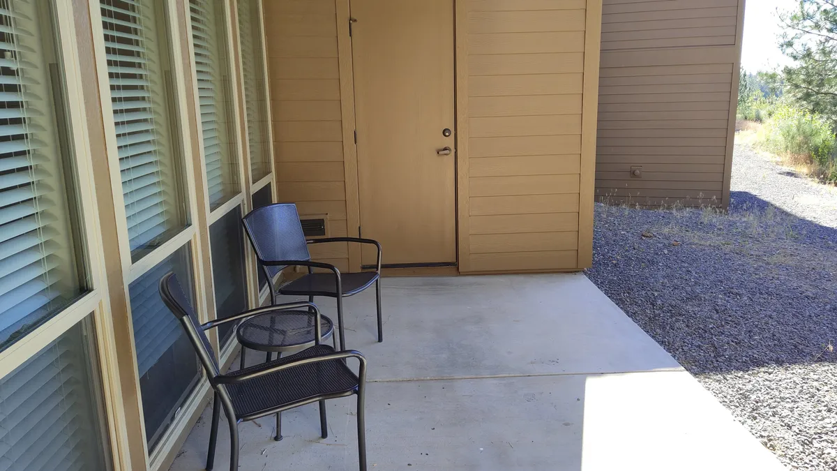 This image showcases an exterior seating area with two black chairs positioned near the front door, framed by large windows and surrounded by natural elements. The setup suggests a cozy space to sit and relax while enjoying the outdoors, echoing the idea of a private, intimate corner.