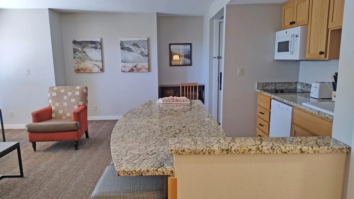 This image features a granite-topped kitchen counter, a bright red accent chair, framed artwork, and modern kitchen appliances. The space feels casual and comfortable, ideal for quick breakfasts or prepping snacks before heading out for activities.