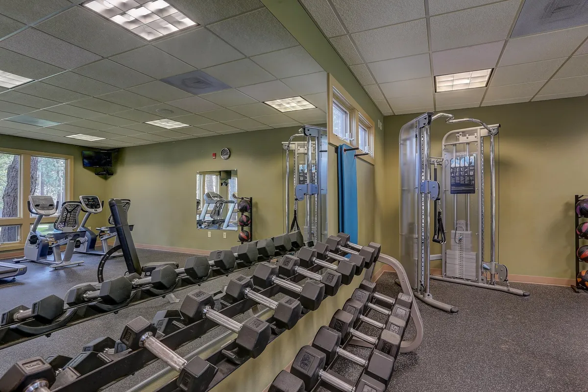 A well-equipped fitness room with a dumbbell rack, various exercise machines, and large mirrors. The space is bright and motivational, designed for solo workouts or group fitness sessions.