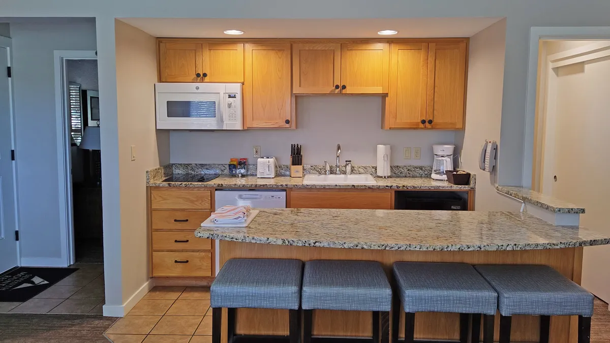 A bright kitchenette scene with a gleaming granite countertop, wooden cabinets, bar stools at the counter, and modern kitchen appliances. The space is set for casual dining or preparing meals together.