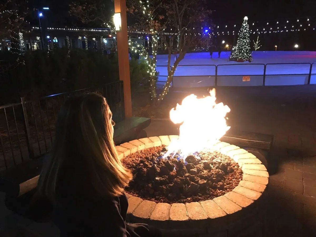 A festive outdoor scene with a fire pit, a person enjoying the warmth, a decorated Christmas tree, and an ice skating rink illuminated by holiday lights. The mood is lively and warm, highlighting the resort’s capacity for winter and holiday activities.