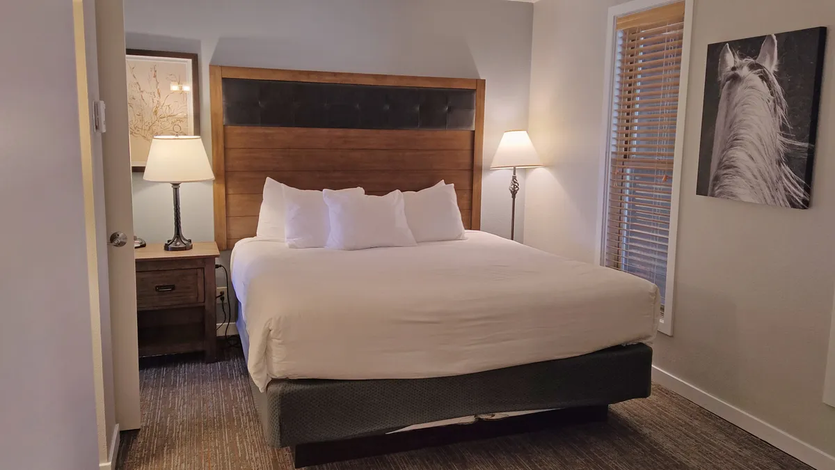 A calm and serene bedroom featuring a plush king bed dressed in soft linens, with a nightstand and warm lamp beside it. Above the bed, a striking horse artwork adds a touch of personality. The space feels airy and inviting, perfect for restful sleep or quiet reading.