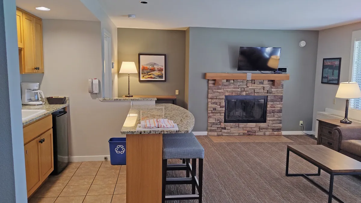 An inviting open-plan living area featuring a kitchen bar with stools, a prominent fireplace, a flatscreen TV, and a comfortable seating area. The kitchen area is equipped with visible appliances and is adjacent to the cozy living zone, all within one seamless space.